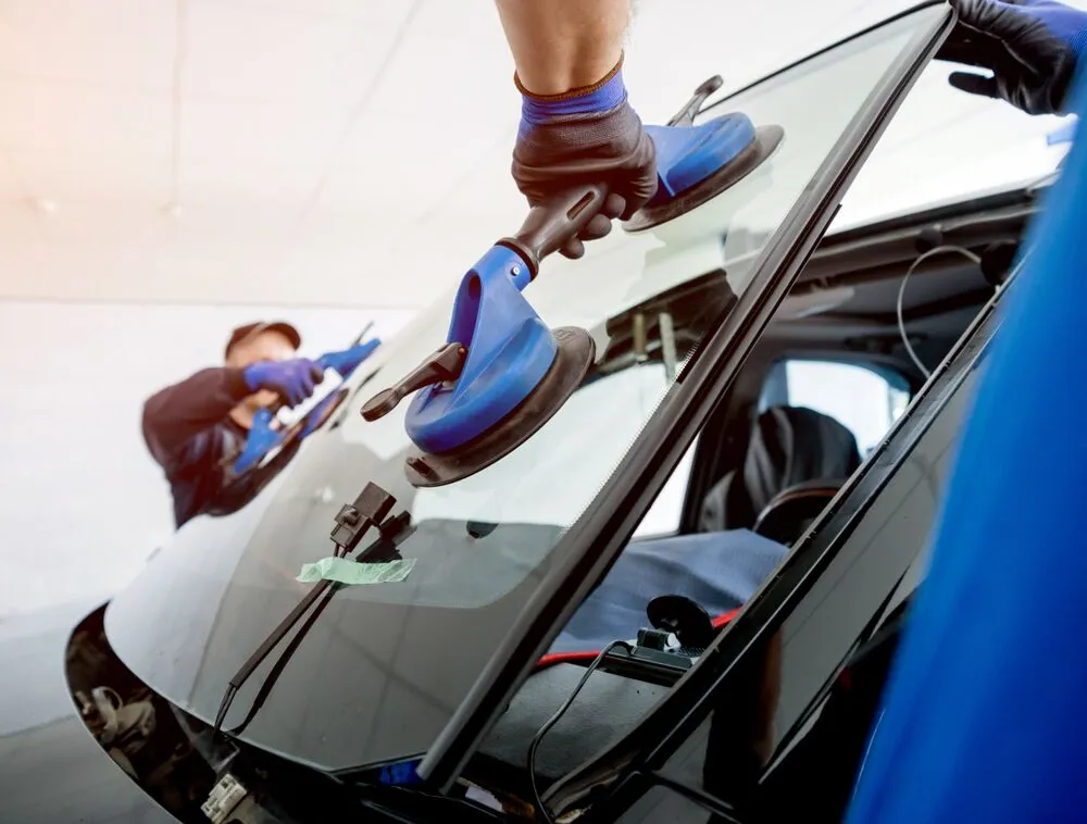 Gloved technician lifting a car windshield into place with blue suction-cup handles
