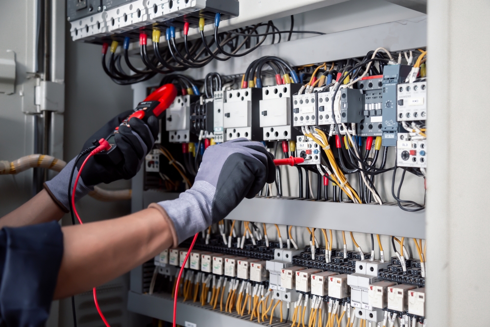 Gloved hands testing an industrial electrical panel with a multimeter probe, among rows of contactors and wired terminals