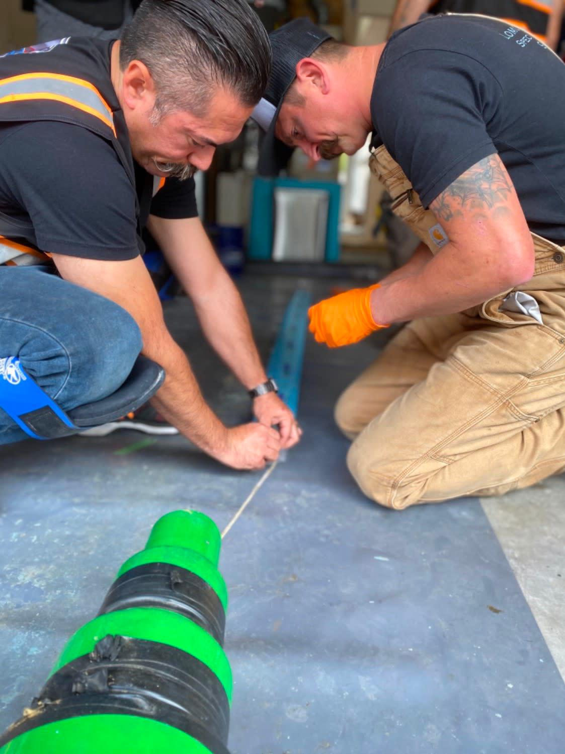 Two workers kneeling on a shop floor, measuring a green and black trenchless pipe liner with a tape measure