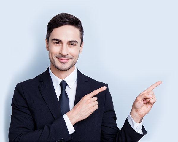 Check this out! Young cheerful successful brunete  lawyer on the pure light blue background is smiling, wearing suit with tie and is pointing on a copyspace with his fingers